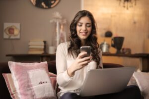 woman sitting on sofa while looking at phone with laptop on lap